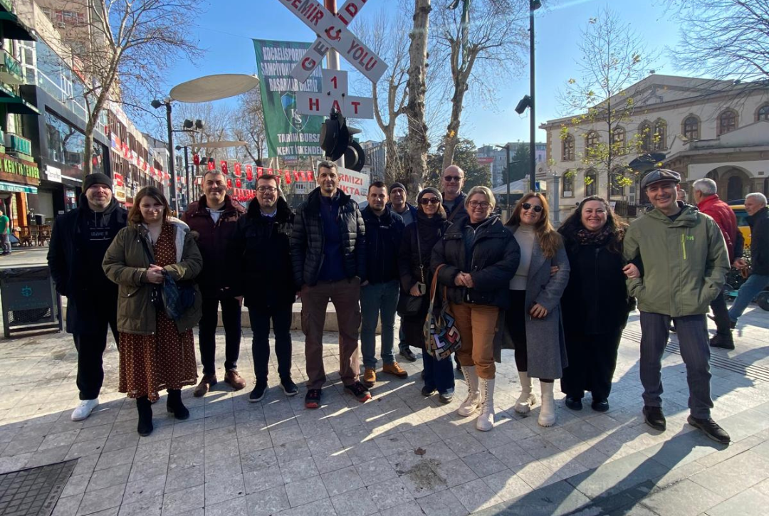 Partners of RISS project smiling in an urban area with a "Demir ve Yolu" sign and trees in the background